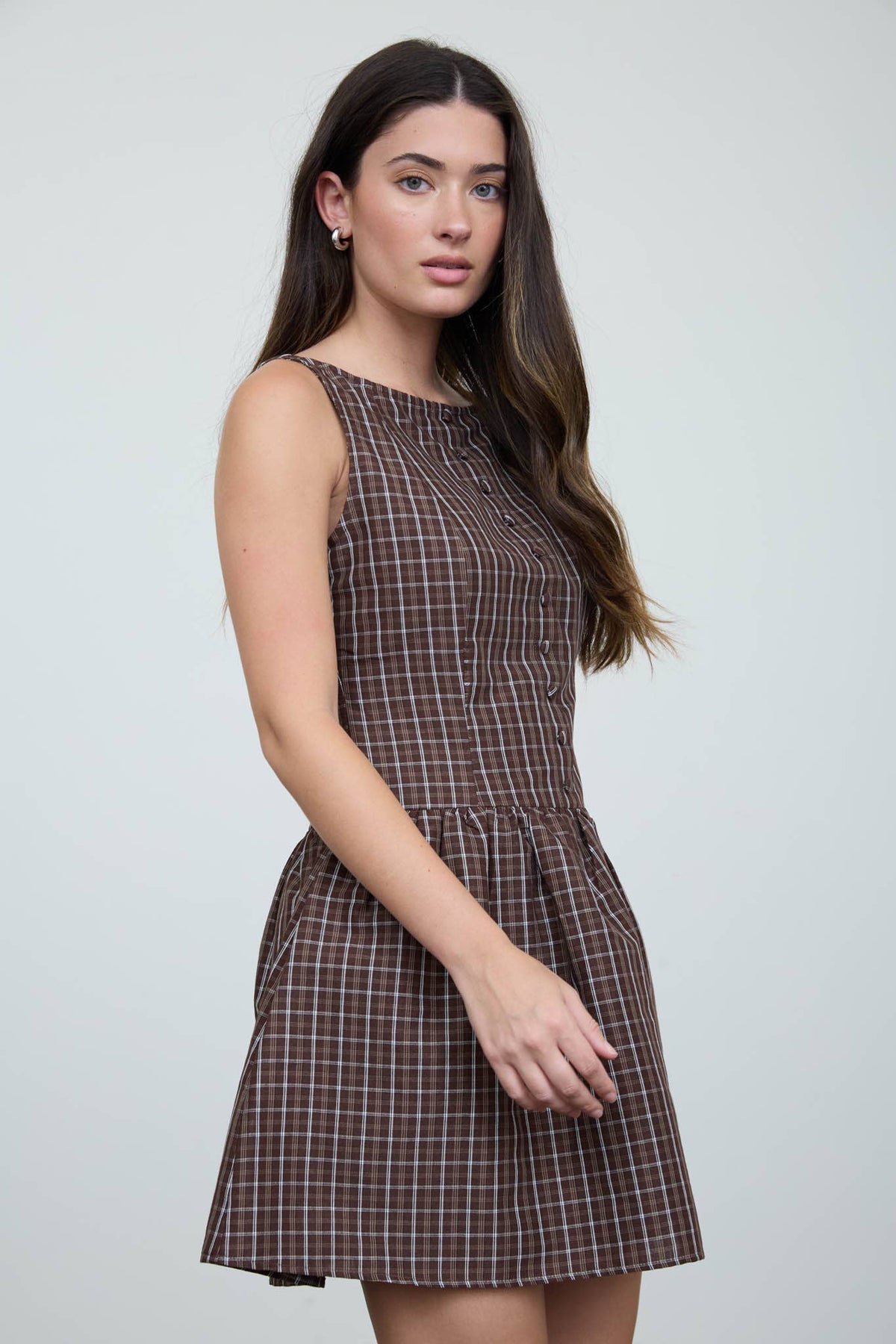 A woman with long brown hair stands against a plain light gray background, looking slightly to the side as she models The Workshop Studio’s Brown Plaid Boat Neck Dress.