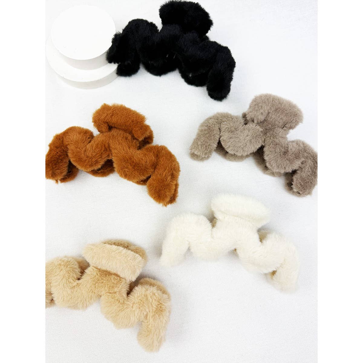 Five Love and Repeat Jumbo Fur Wavy Hair Claw Clips in black, brown, gray, cream, and beige are arranged on a white surface, with circular white props visible in the top left corner.