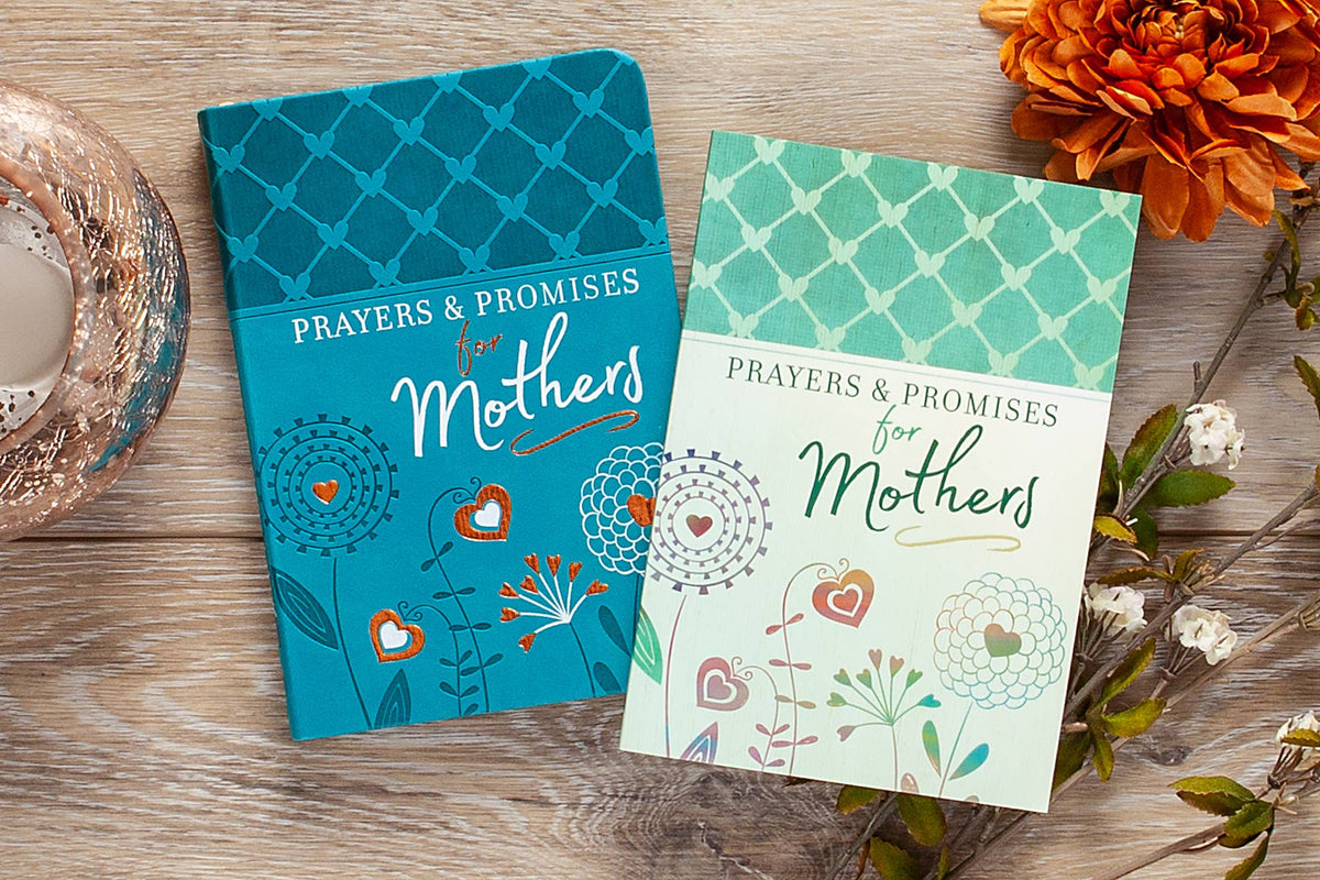 A Prayers & Promises for Mothers - Green book by BroadStreet Publishing Group, LLC and a blue edition are placed on a wooden surface, with flowers and a candle nearby.