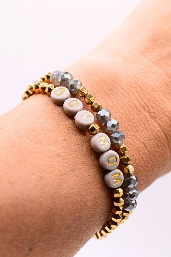 A wrist wearing the Boy Mom Bracelet Set by Two and Crew - Faith Based Jewelry & Gifts; one bracelet features gold and silver beads with white letter beads spelling “BOY MOM.”.