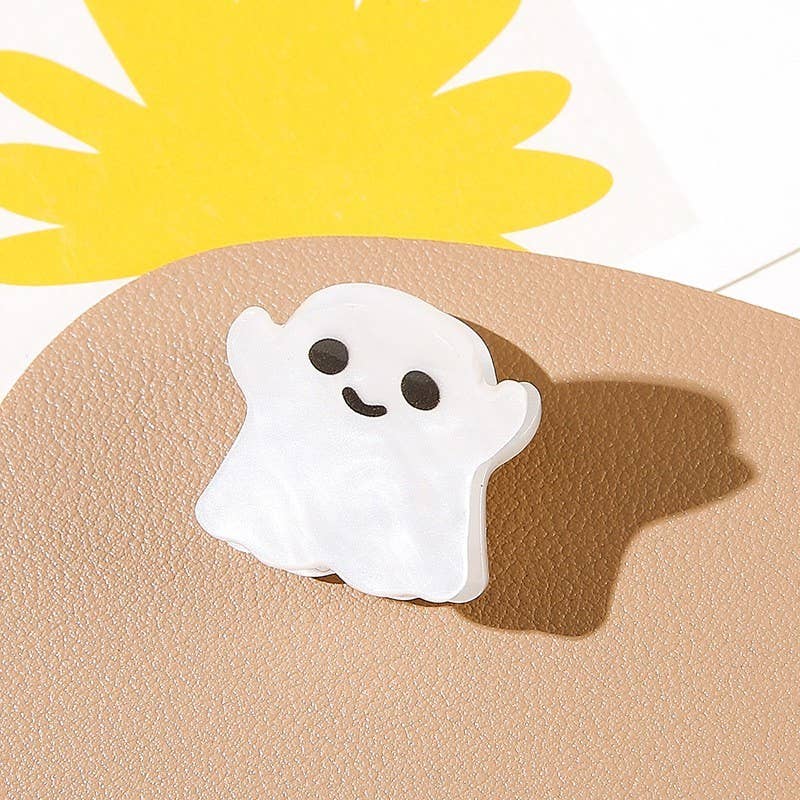 A small, smiling ghost-shaped mini hair clip from the Halloween Ghost Theme Mini Hair Clips 4-Pack by JOSSLYN by wall to wall, featuring black eyes and a mouth, is displayed on a light brown textured surface with a yellow abstract shape partially visible in the background.