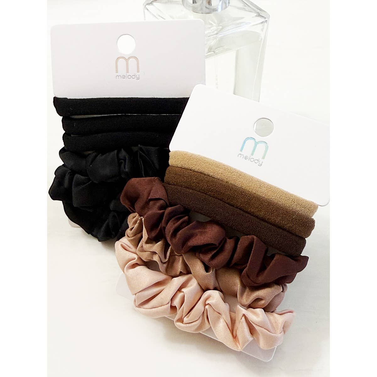 Two packs of the Love and Repeat Assorted Hair Tie & Scrunchie Set, featuring black, brown, and beige shades, are displayed upright on white cardboard holders with branded packaging. A clear bottle is partially visible in the background.