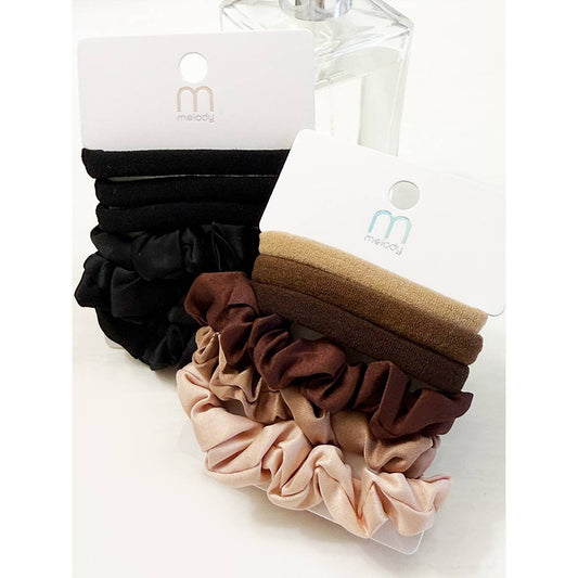Two packs of the Love and Repeat Assorted Hair Tie & Scrunchie Set, featuring black, brown, and beige shades, are displayed upright on white cardboard holders with branded packaging. A clear bottle is partially visible in the background.