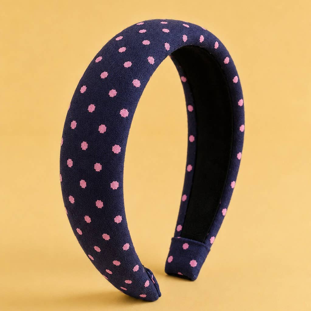 The JOSSLYN by wall to wall Padded Polka Dot Fabric Headband, featuring a navy blue base with pink polka dots, is displayed standing upright against a plain yellow background.