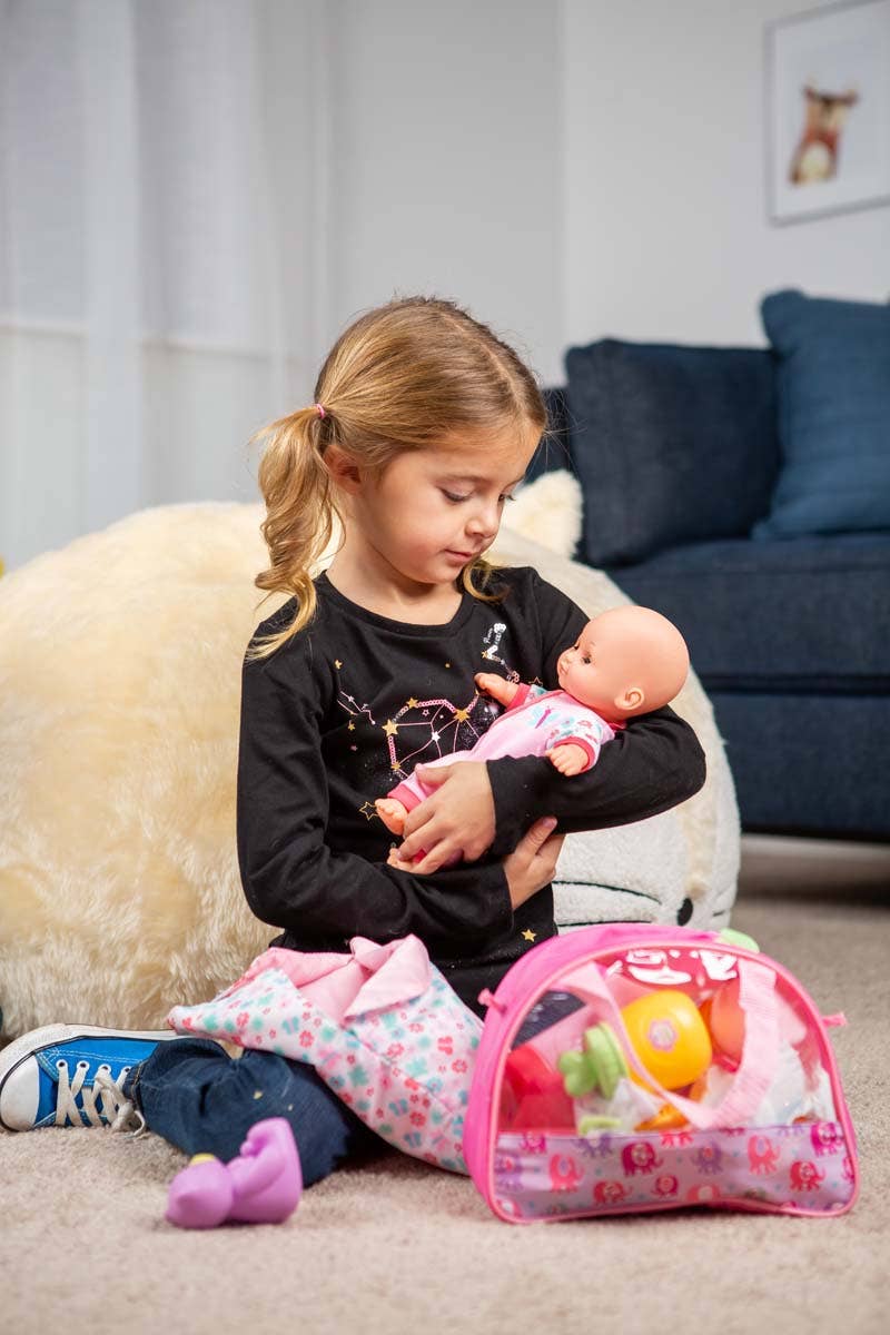 A young girl sits on the floor, cradling a baby doll from the Toysmith My Sweet Baby Deluxe Baby Ensemble 12-Piece Doll Playset. She wears a black star-patterned shirt and colorful pants. Nearby, a pink toy bag and plush pillow are surrounded by doll accessories from the set—perfect for imaginative pretend play. In the background, a dark blue couch completes the cozy scene.
