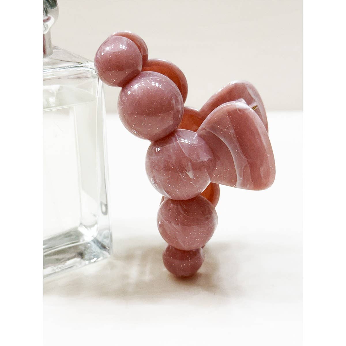 The Large Marbled Hair Claw by Love and Repeat, featuring a pink, glossy finish with a geometric, bubbly design, rests against a clear glass bottle on a white surface.