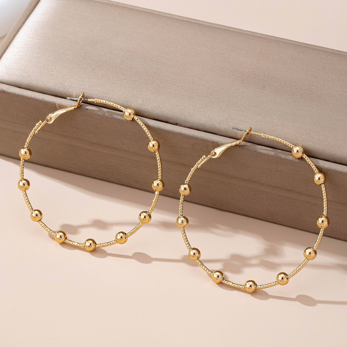 Gold Ball Beaded Hoop Earrings by JOSSLYN by wall to wall are displayed on a beige box, with their evenly spaced gold beads casting soft shadows on a light surface.