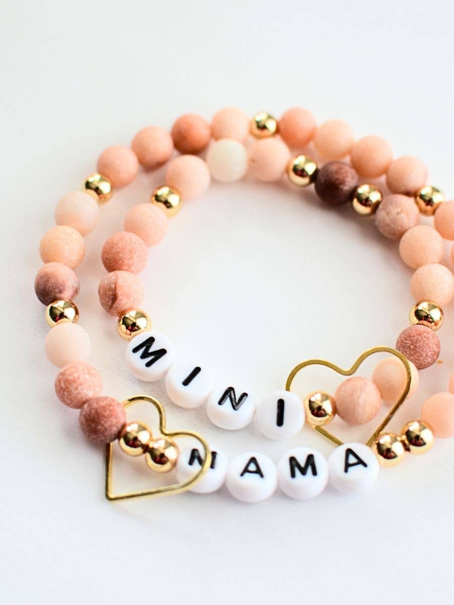 The Mama Mini Bracelet Set by Two and Crew - Faith Based Jewelry & Gifts features two beaded bracelets adorned with pink, brown, and gold beads. Each bracelet includes a gold heart charm and white letter beads that spell out “MINI” and “MAMA.” The bracelets are displayed in a spiral arrangement on a white background.