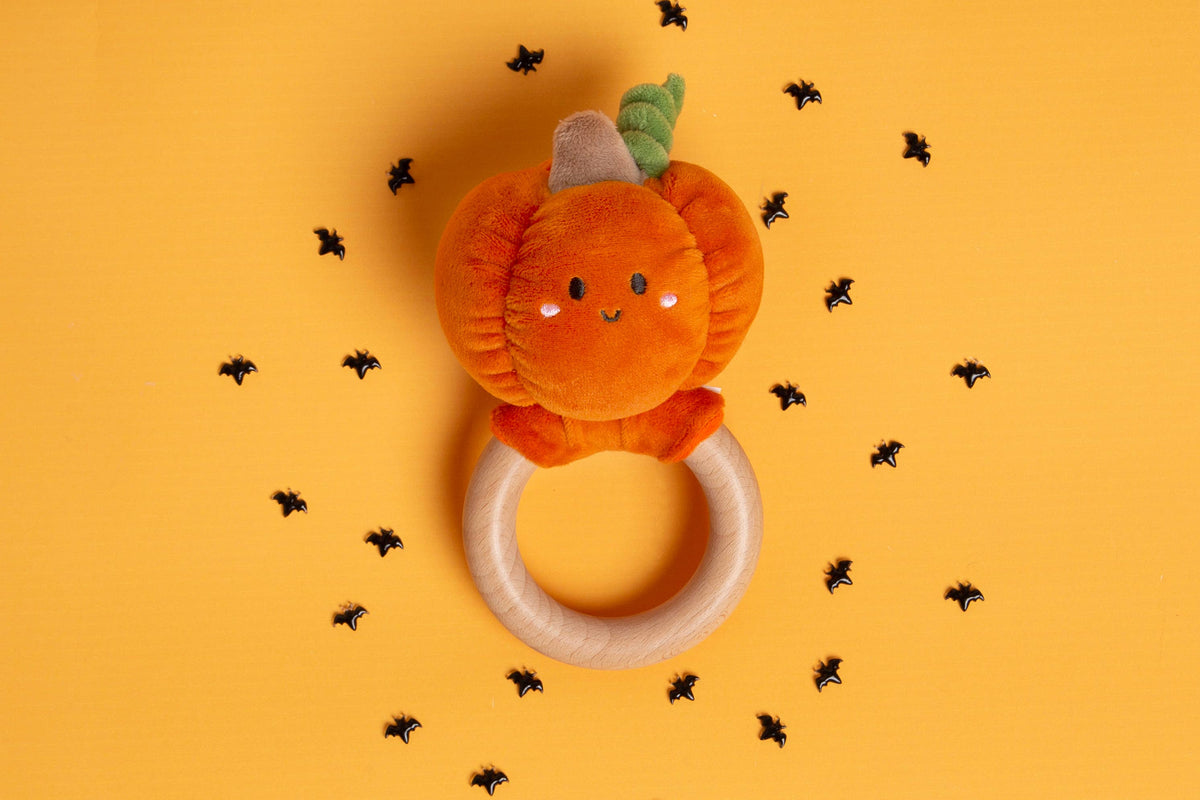 Pearhead Pumpkin Baby Wooden Teether Toy for Halloween and Fall, featuring a plush pumpkin on a smooth wooden ring surrounded by bat-shaped confetti on an orange background.