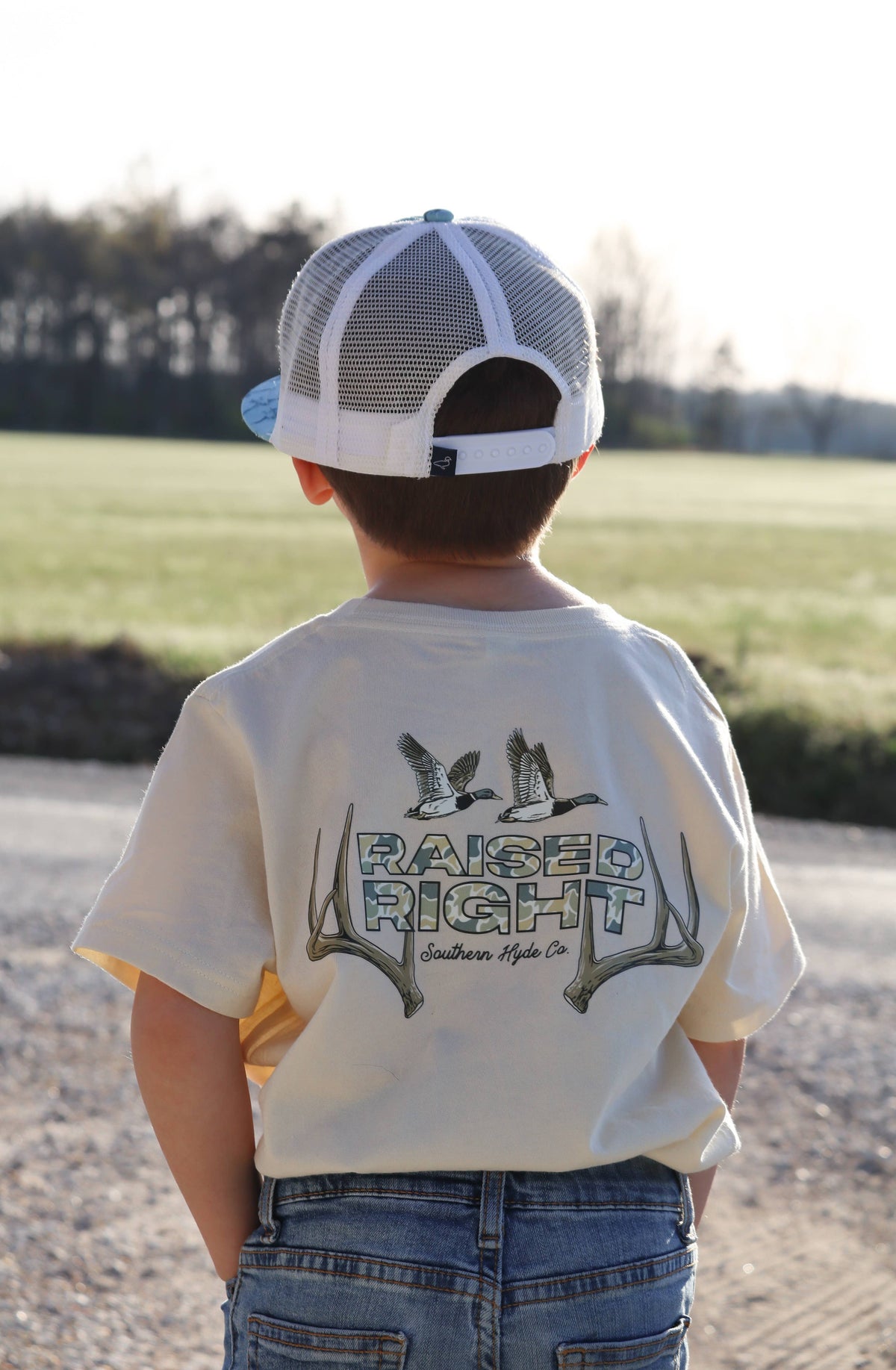 A young child viewed from behind wearing a white mesh baseball cap and a cream-colored Raised Right Tee with a graphic of two ducks flying over deer antlers.