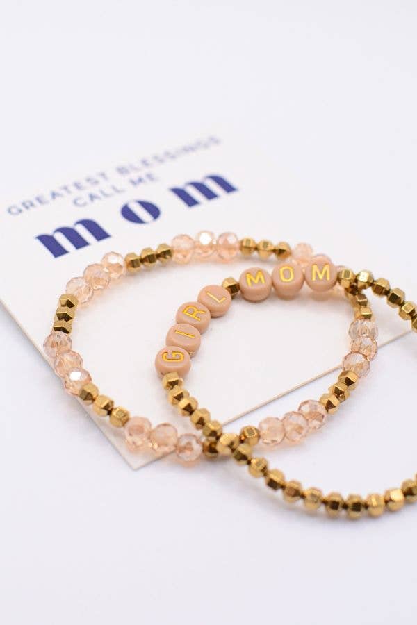 The Girl Mom Bracelet Set by Two and Crew - Faith Based Jewelry & Gifts features two beaded bracelets—one with gold and clear beads, and the other with letter beads spelling out “GIRL MOM”—displayed on a card that says “GREATEST BLESSINGS CALL ME mom” in blue and gold letters.