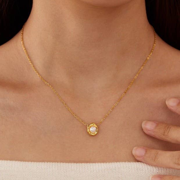 A close-up of a woman wearing the JOSSLYN by wall to wall Tarnish Free Pearl Necklace, featuring a delicate gold chain with a round white pearl pendant. Her hand gently rests on her collarbone as she is dressed in a light, off-shoulder top.