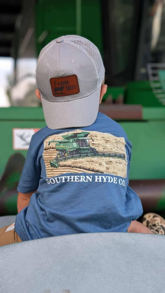 A young child seen from behind wears a gray baseball cap and the Southern Hyde Co. Grain Train Tee, a blue t-shirt with a graphic of a combine harvester in a field.