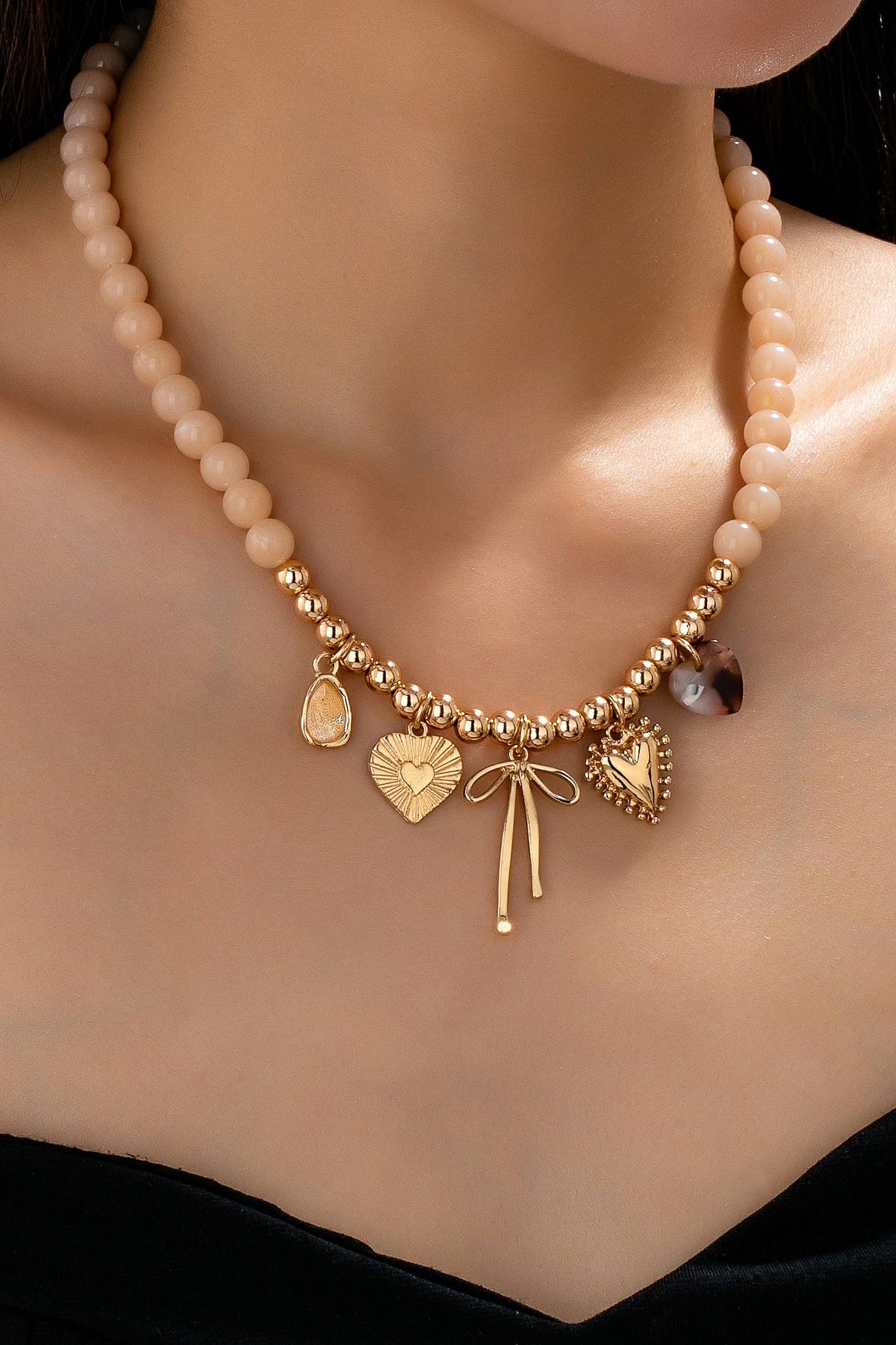 A close-up of a woman wearing the JOSSLYN by wall to wall Beige Beaded Bow Heart Rhinestone Charm Necklace, featuring gold heart, bow, and teardrop charms, against her bare skin and a black top.
