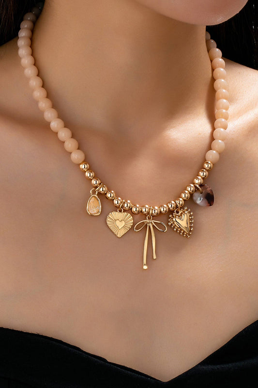 A close-up of a woman wearing the JOSSLYN by wall to wall Beige Beaded Bow Heart Rhinestone Charm Necklace, featuring gold heart, bow, and teardrop charms, against her bare skin and a black top.