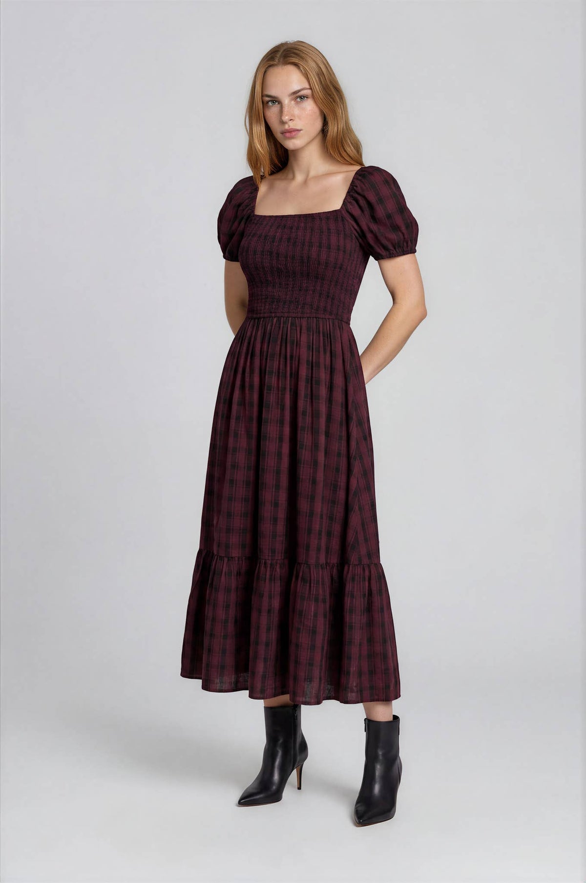 A person wears a Blu Pepper Burgundy Plaid Midi Dress in burgundy and black with a square neckline, puffed short sleeves, and a shirred bodice, paired with black heeled ankle boots.