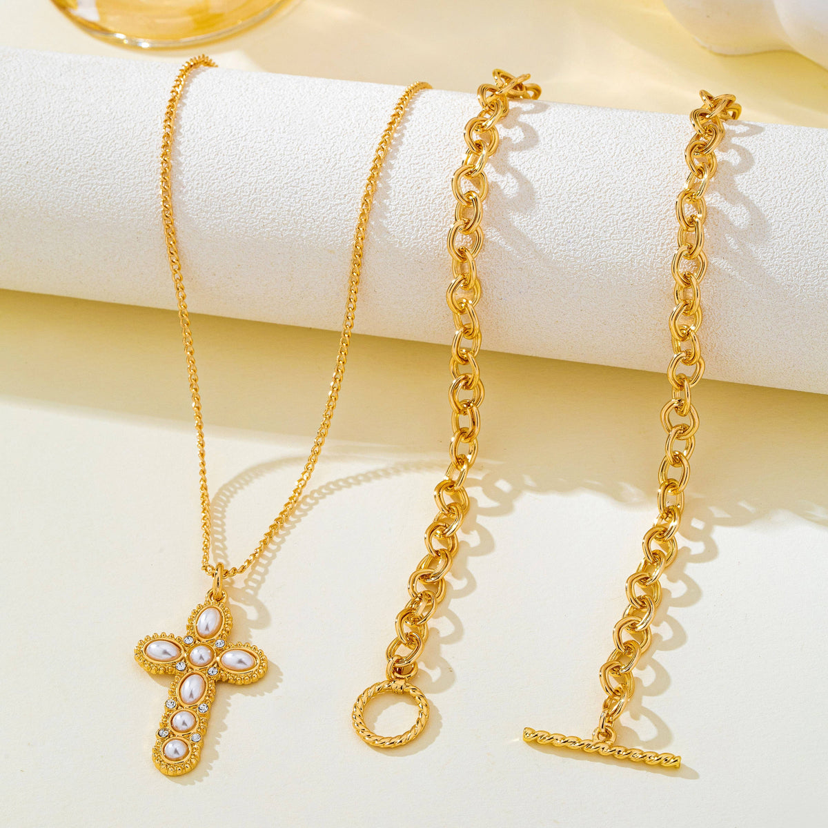 JOSSLYN by wall to wall Pearl Cross Layered Necklace Set displayed on a cylindrical stand, featuring a gold-tone cross pendant with white pearls and crystals alongside a thicker gold-tone chain with a toggle clasp.