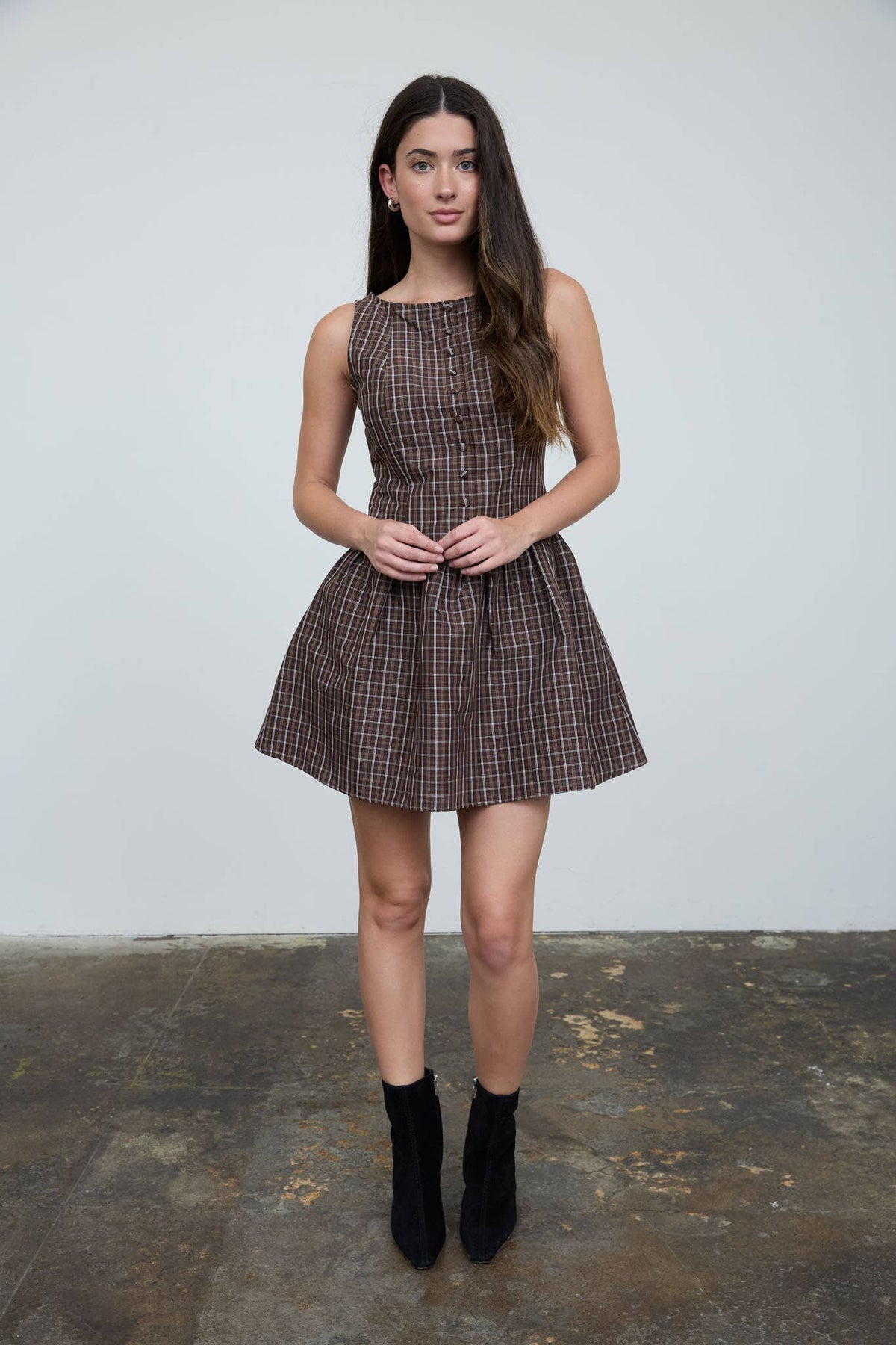 A woman stands indoors on a concrete floor, facing the camera with her hands clasped in front of her, wearing The Workshop Studio’s Brown Plaid Boat Neck Dress with a pleated skirt and black ankle boots.