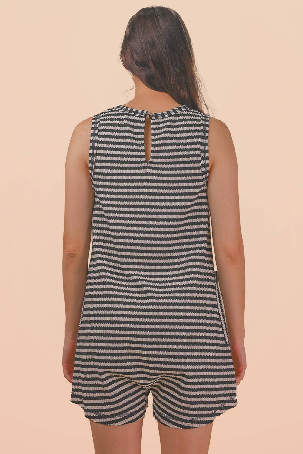 A woman with long brown hair is seen from behind, modeling the VERY J Striped Knit Casual Romper—a sleeveless, black and white striped piece—against a plain peach background.