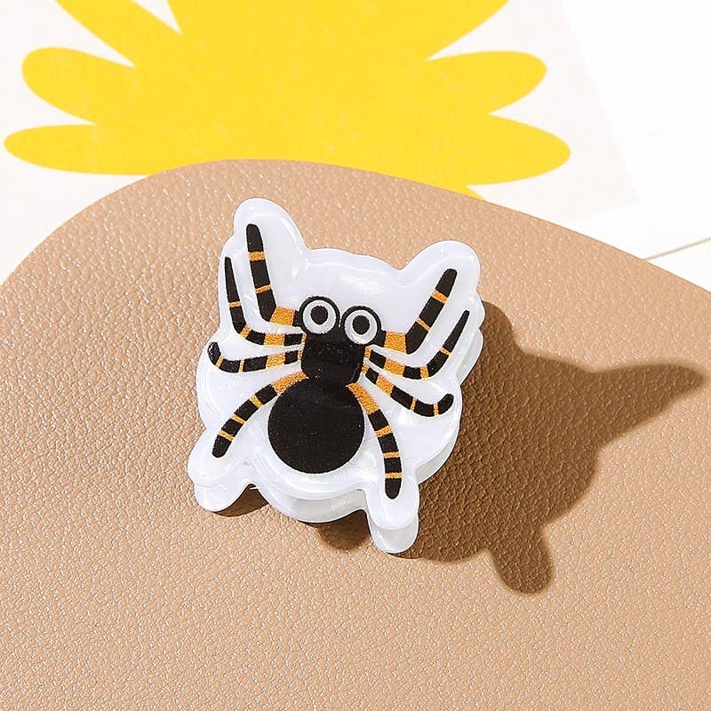 A mini hair clip from the JOSSLYN by wall to wall Halloween Ghost Theme 4-Pack, featuring a small cartoon spider with black and orange stripes, big googly eyes, and shown on a tan surface next to a yellow flower illustration.
