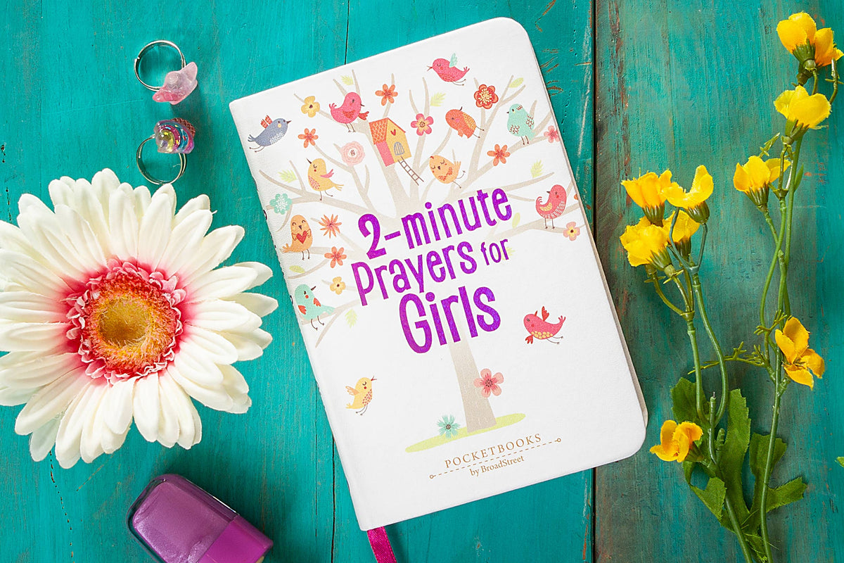 The 2-Minute Prayers for Girls Book by BroadStreet Publishing Group, LLC rests on a turquoise surface next to yellow flowers, a daisy, rings, and pink nail polish. Its cover showcases colorful birds perched on a tree illustration.