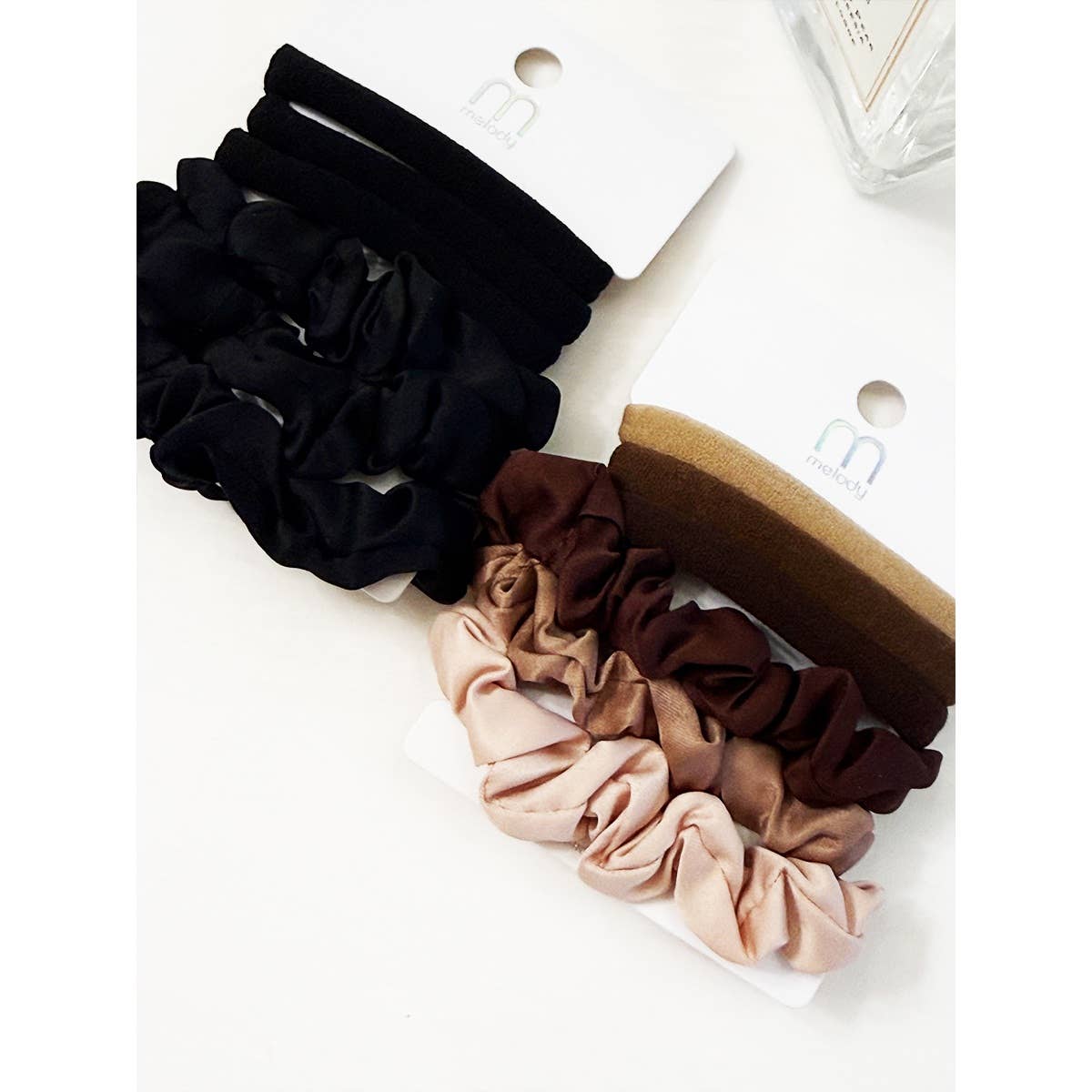 Eight scrunchies from the Love and Repeat Assorted Hair Tie & Scrunchie Set—four in black and four in brown tones—are displayed on white logo cards, arranged on a white surface beside a clear glass container.
