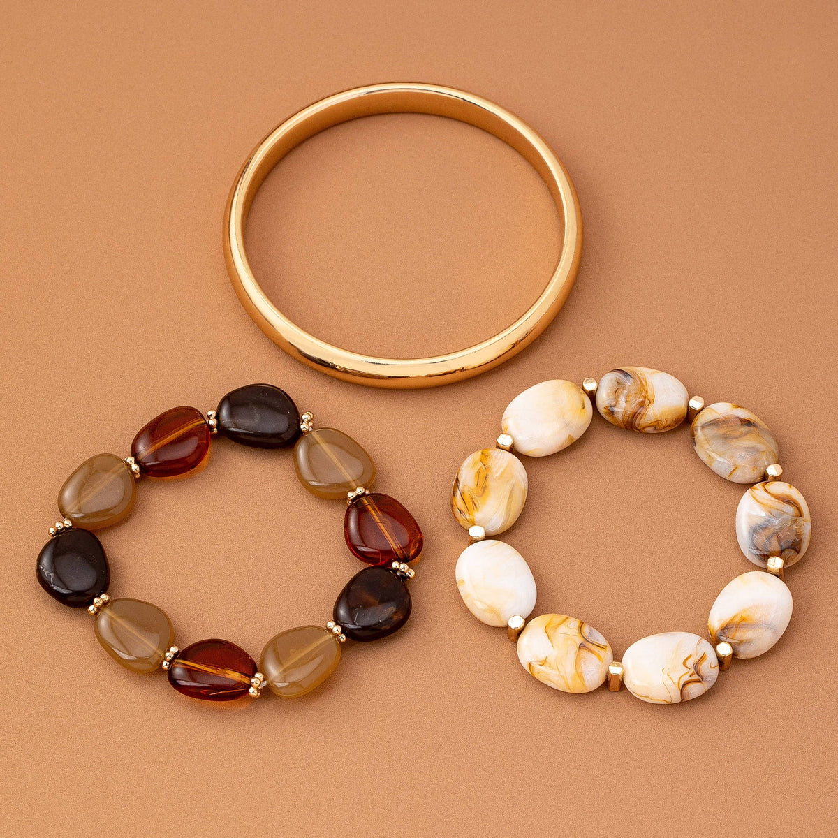 Three bracelets from the JOSSLYN by wall to wall Boho Gold Bracelet Set are displayed on a tan background: a gold bangle, a beaded bracelet with brown and amber stones, and a beaded bracelet with white and beige marbled stones.