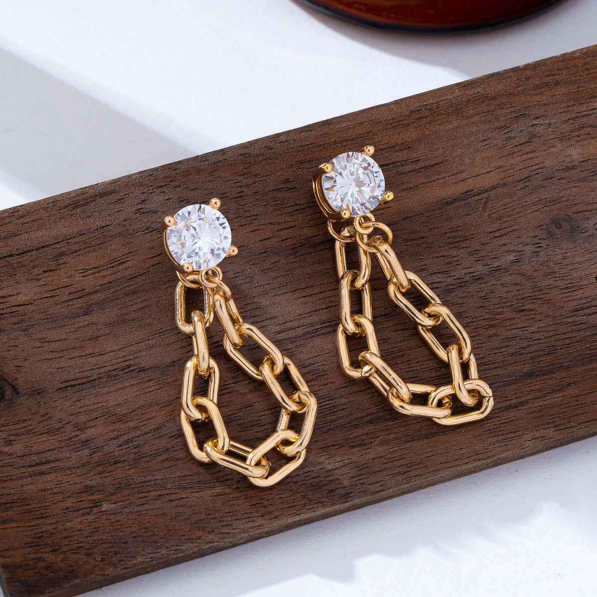 Stud Hoop and Drop Link Earrings by JOSSLYN by wall to wall, featuring gold chain links with a round, clear gemstone stud, displayed on a wooden surface.