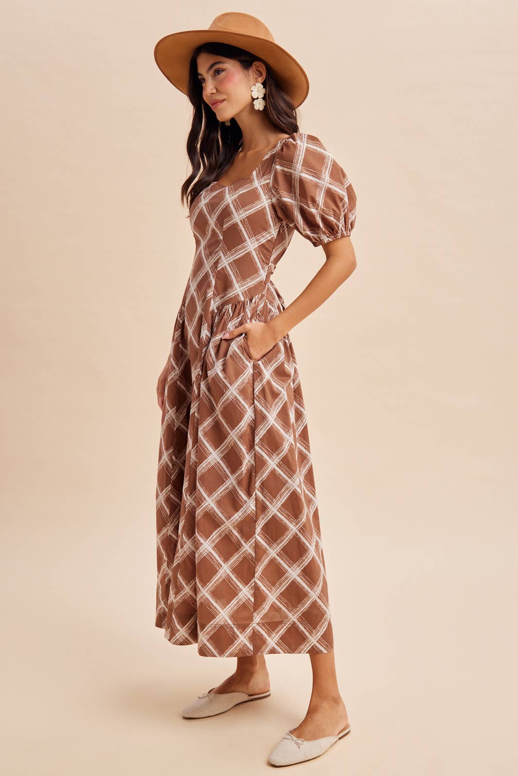 A person wearing an In Loom Plaid Square Neck Maxi Dress in brown plaid with short puff sleeves, a square neckline, and side pockets, paired with a wide-brim hat and slip-on shoes.