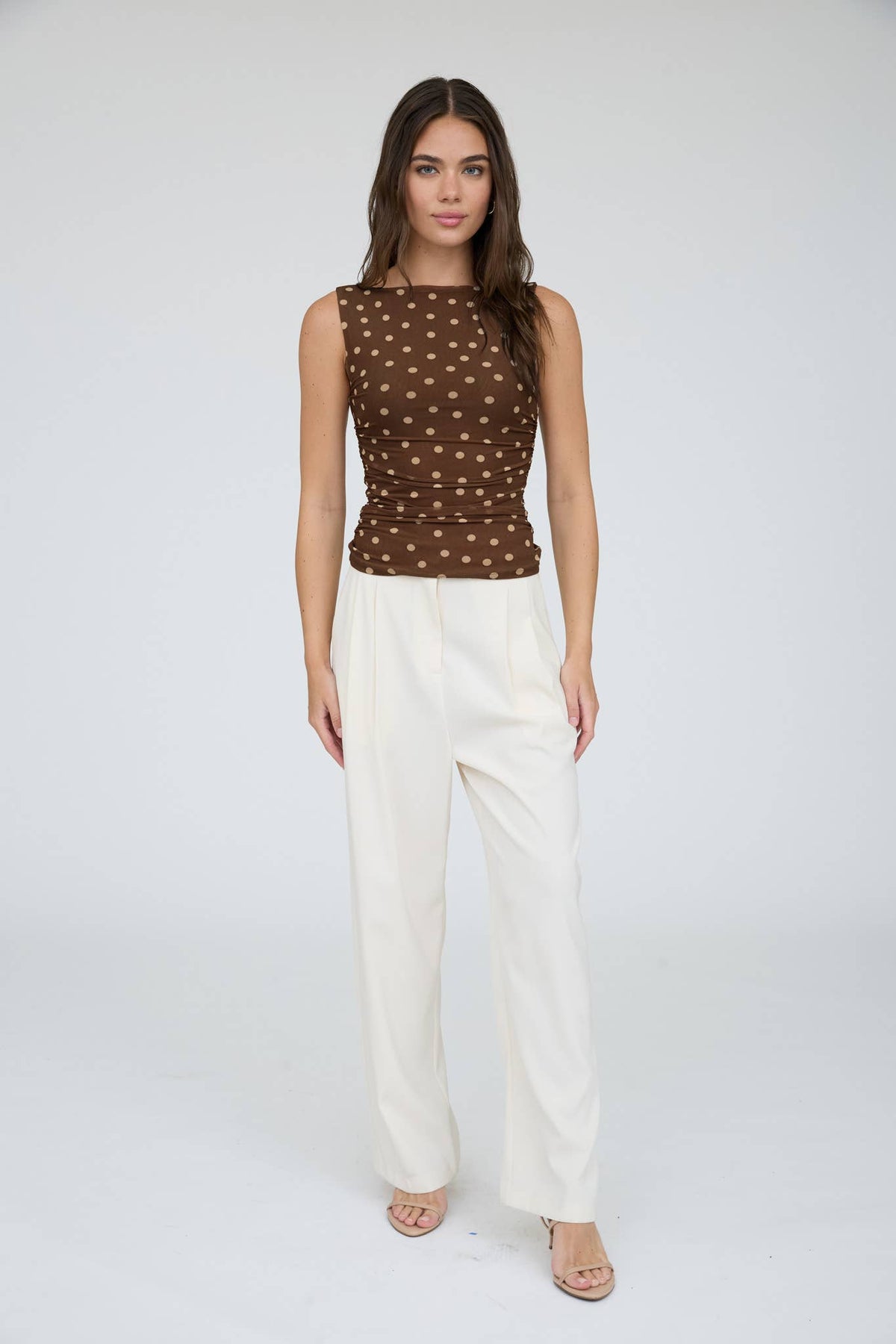 A woman stands in a studio against a plain light background wearing The Workshop Studio’s Brown Polka Dot Boat Neck Top, styled with white high-waisted wide-leg pants and nude open-toe heels.