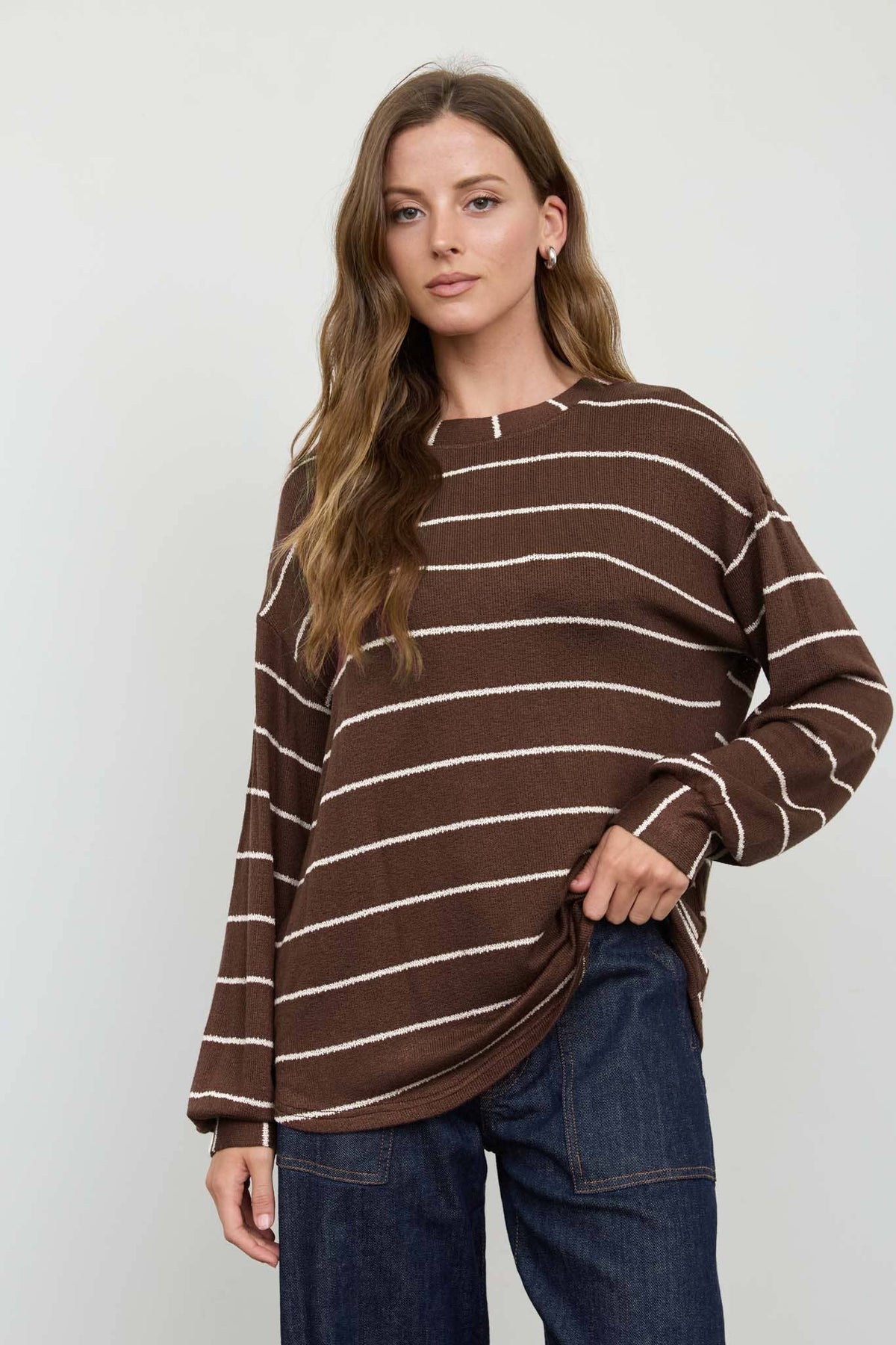 A woman with long wavy hair wears the Blu Pepper Brown Stripe Cuffed Knit Top, paired with dark blue jeans, as she stands against a plain light background.