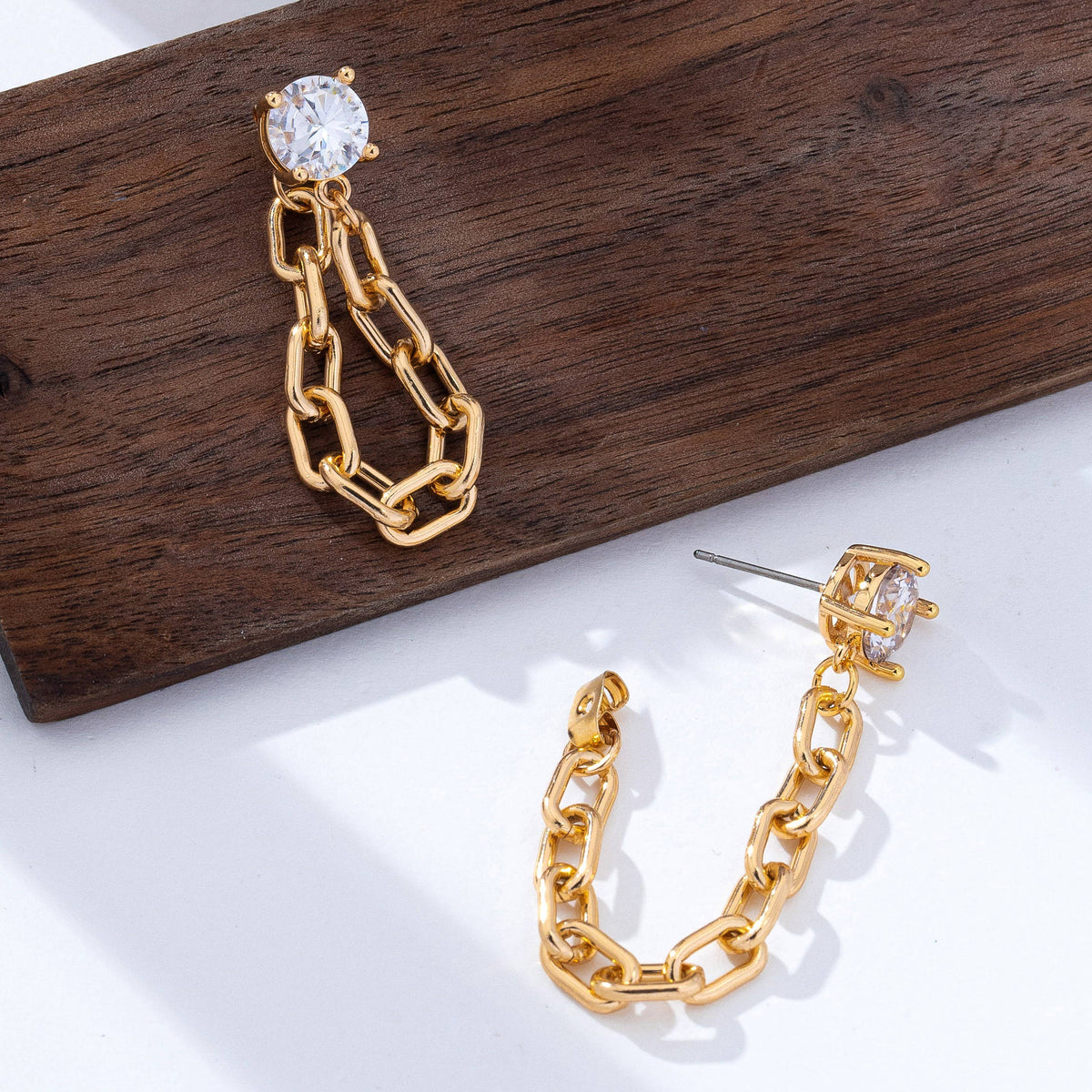 Stud Hoop and Drop Link Earrings from JOSSLYN by wall to wall, featuring gold chain links and a round clear gemstone stud, displayed on a wooden board and white surface with natural light and soft shadows.