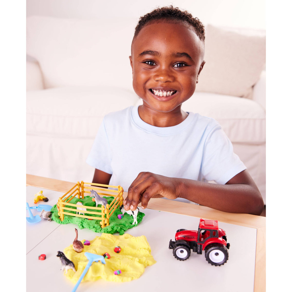 A smiling child enjoys imaginative play with the Faber-Castell Creativity for Kids Sensory Pack Farm On the Go Sensory Play Set, creating a yellow playdough farm scene and playing with a red toy tractor on a table in a bright living room.
