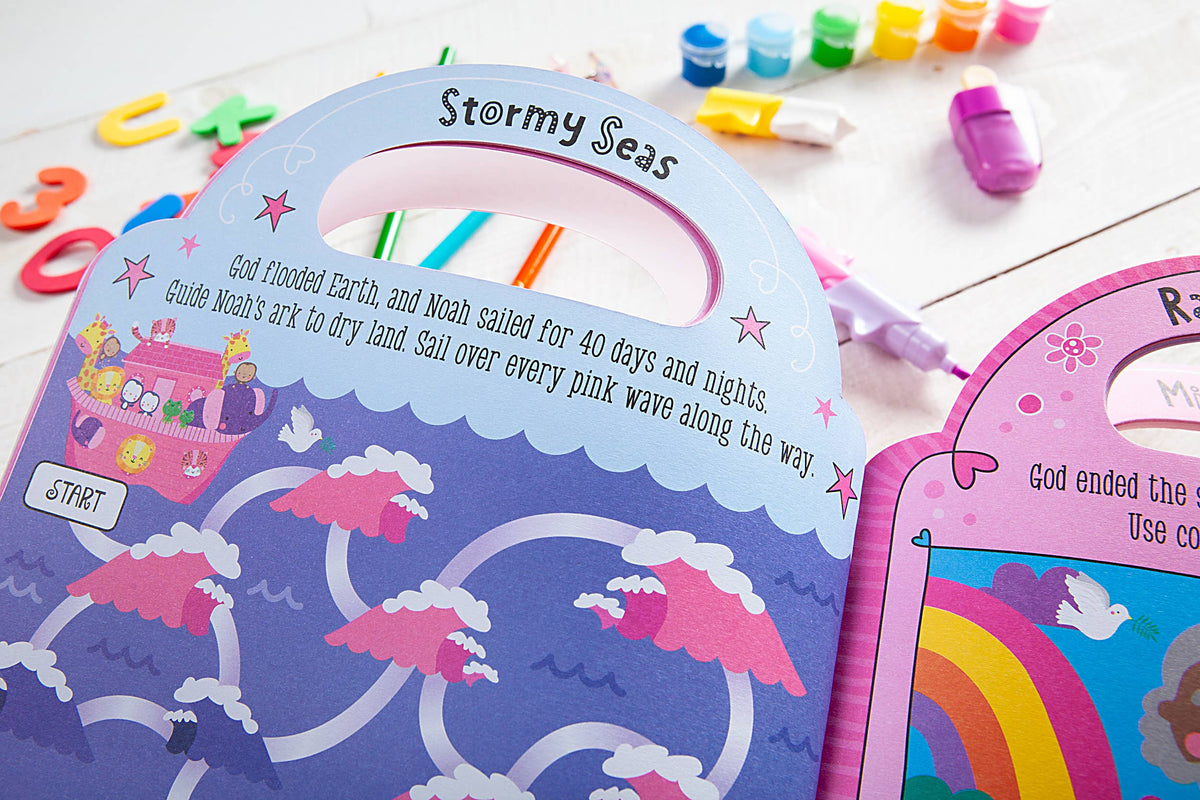 The My Pretty Pink Bible Activity Book by BroadStreet Publishing Group, LLC is opened to a colorful Noah’s Ark page featuring vibrant waves, fun text, and an illustrated ark. Markers, paints, and letter magnets are scattered across the white table in the background.