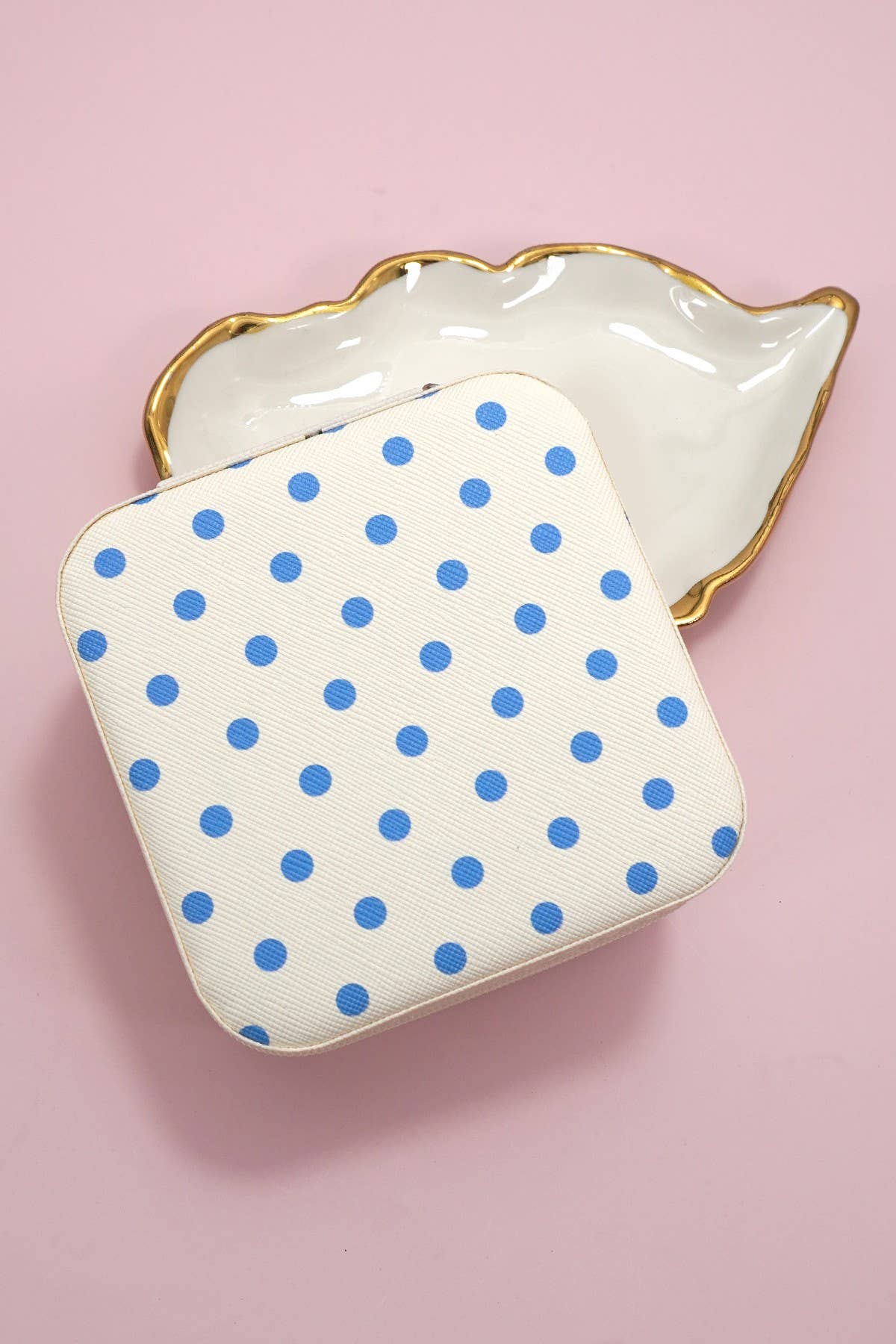 A white, square JOSSLYN by wall to wall Jewelry Organizer Travel Box Case with blue polka dots sits on a pink surface, partly covering a shiny, gold-edged seashell-shaped dish.