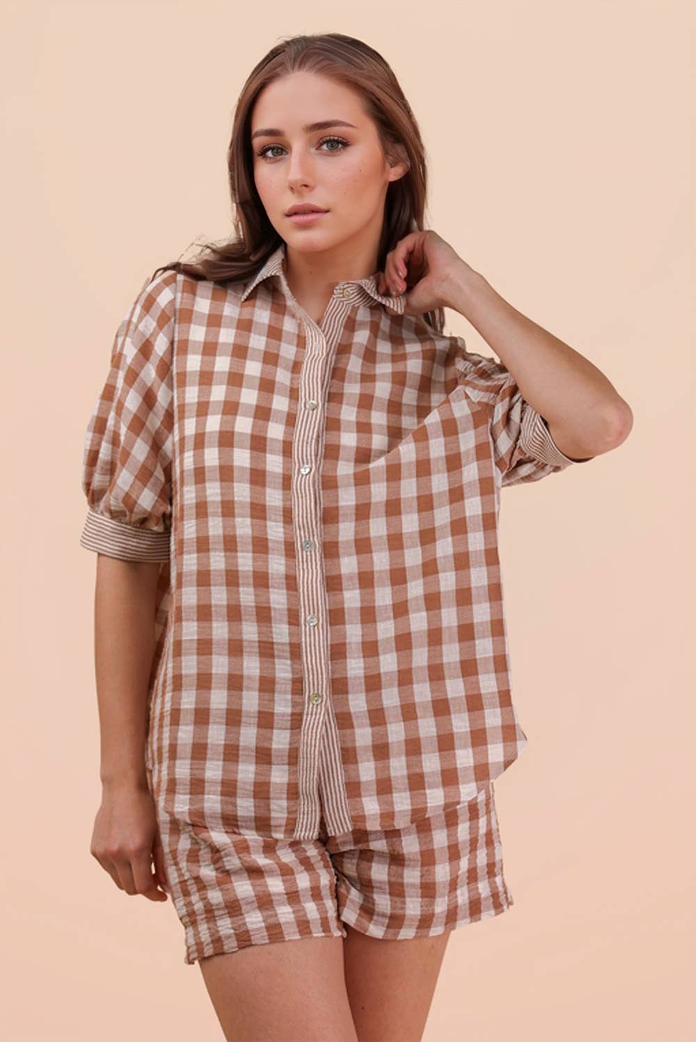 A woman models the VERY J Checkered Shirt Top & Shorts Set, featuring a brown and white checkered button-up shirt and matching shorts, as she stands against a plain beige background and looks at the camera with a neutral expression.