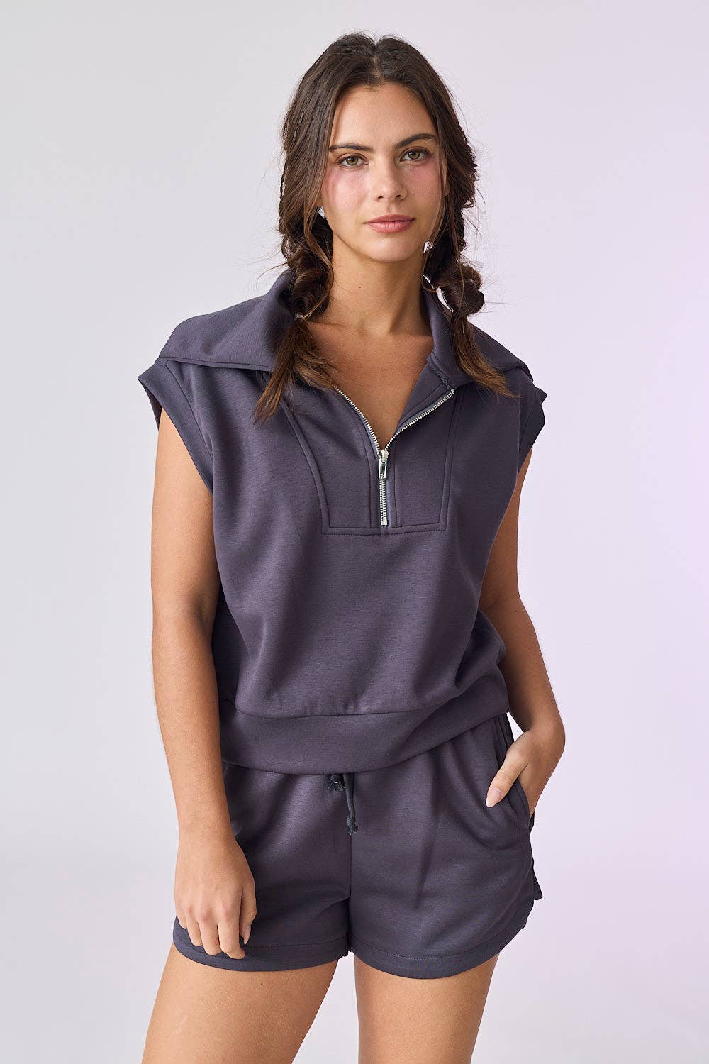 A woman with braided hair wears the Papermoon Blake Half Zip Top and Shorts Set—a sleeveless, zip-collared dark gray ensemble—standing with one hand in her pocket against a plain light background.