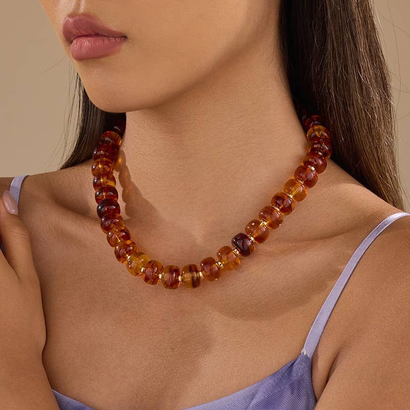 A person wearing a JOSSLYN by wall to wall Chunky Amber-Tone Barrel Bead Necklace, featuring translucent, honey-colored barrel-shaped beads separated by small gold spacers.