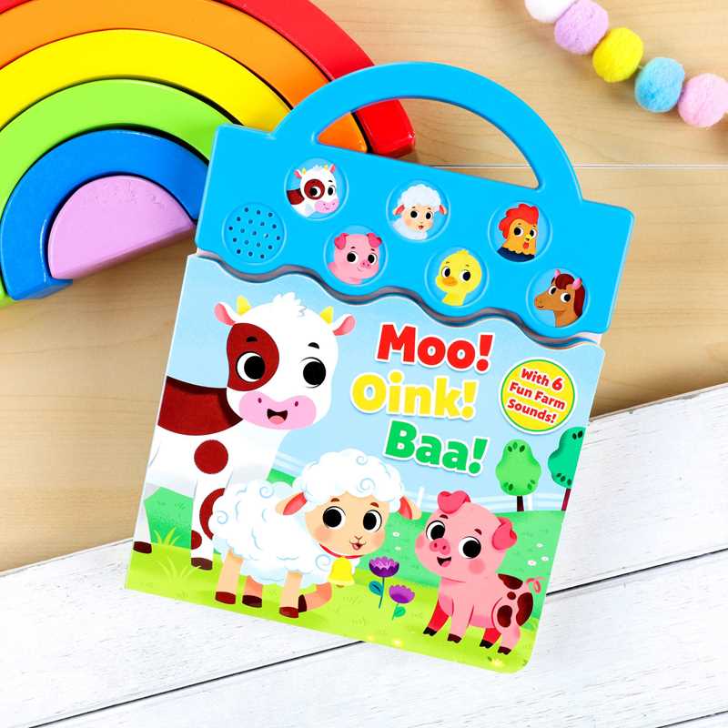 The Moo, Oink, Baa! Book by Simon & Schuster, featuring illustrated cow, sheep, and pig on the cover, rests on a wooden surface beside a rainbow toy and pastel bead garland.