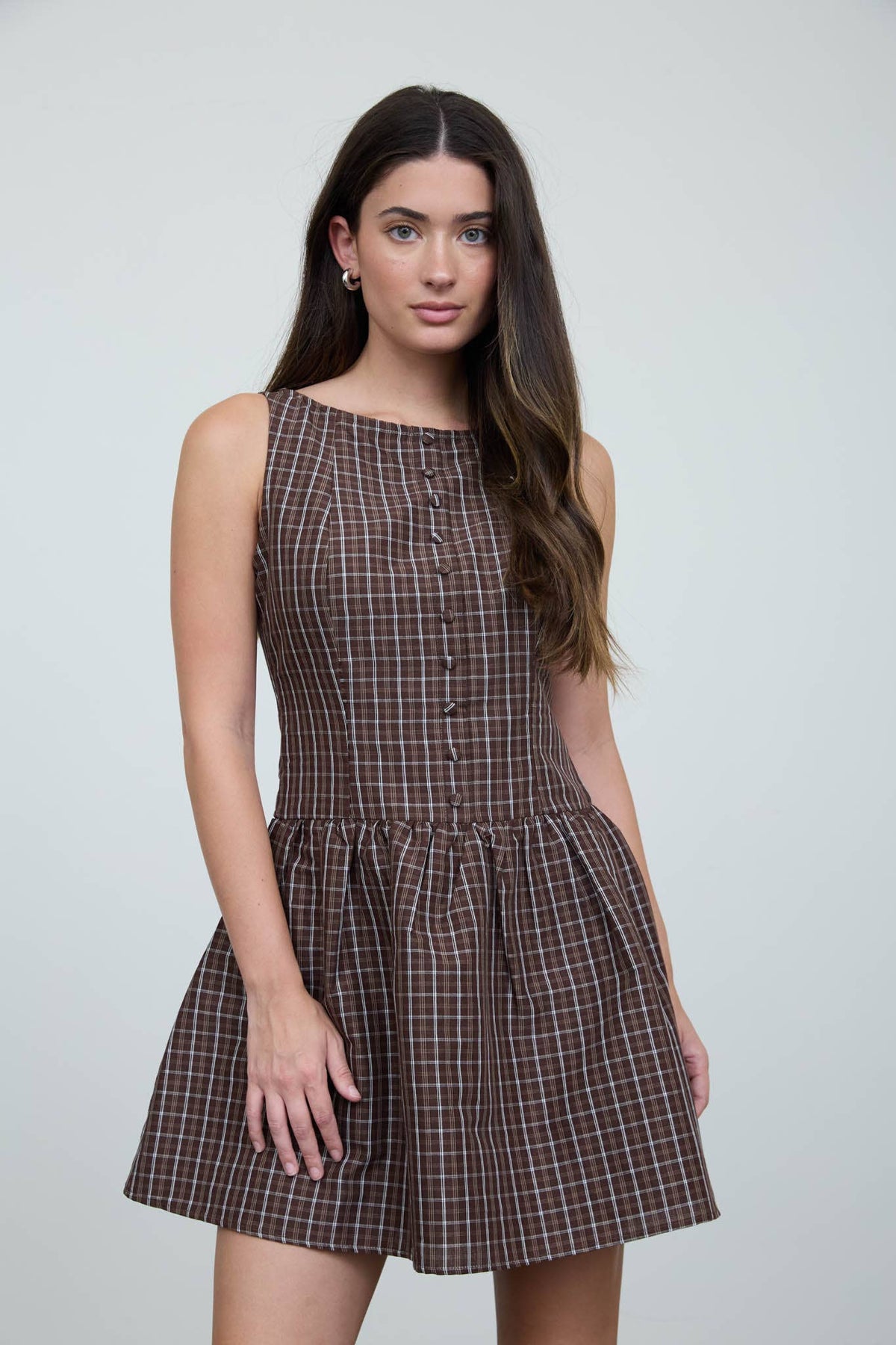 A woman wearing The Workshop Studio’s Brown Plaid Boat Neck Dress, a sleeveless design with front buttons, stands against a plain light background and looks at the camera with one hand by her side.