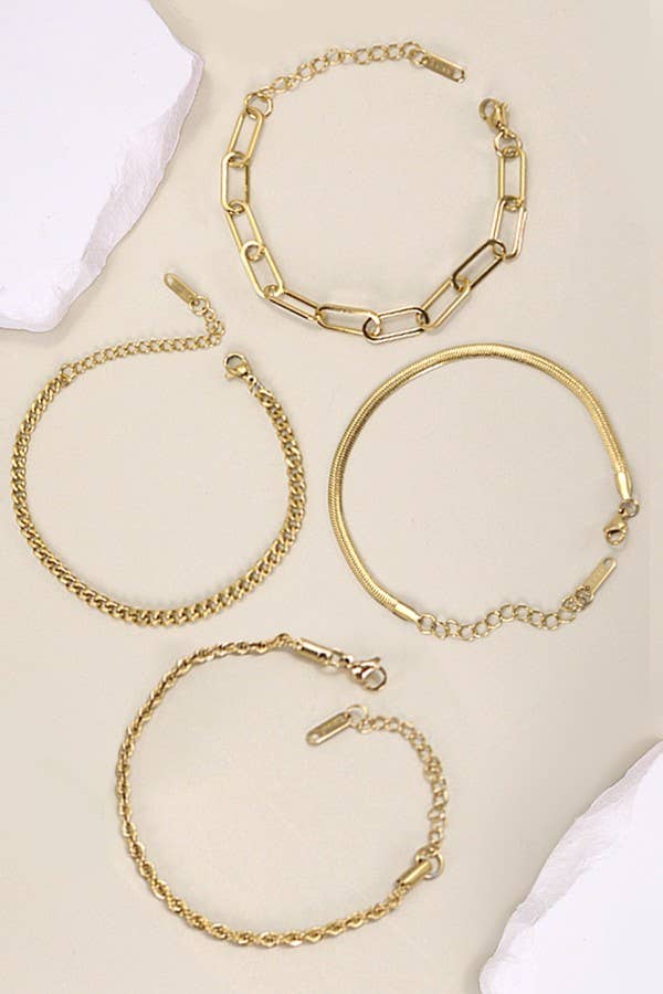 Four Dainty Gold Bracelets from JOSSLYN by wall to wall—each featuring a unique design including large links, a flat snake chain, a rope chain, and a classic curb chain—are displayed on a beige surface accented with white stones.