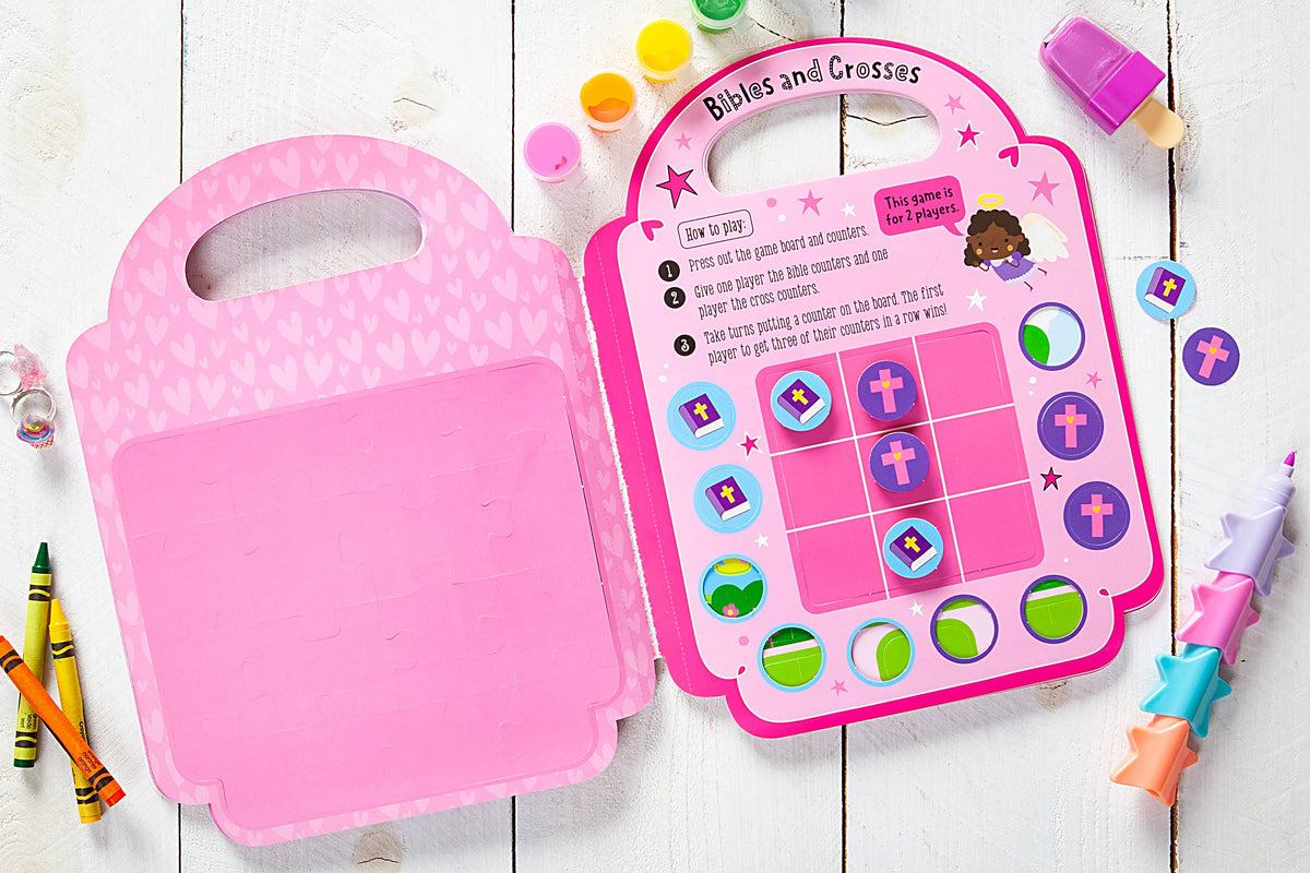 A copy of My Pretty Pink Bible Activity Book by BroadStreet Publishing Group, LLC is open on a white wooden table, surrounded by crayons, paint pots, and a unicorn pen. Colorful tokens shaped like crosses and Bibles are arranged on the activity pages.