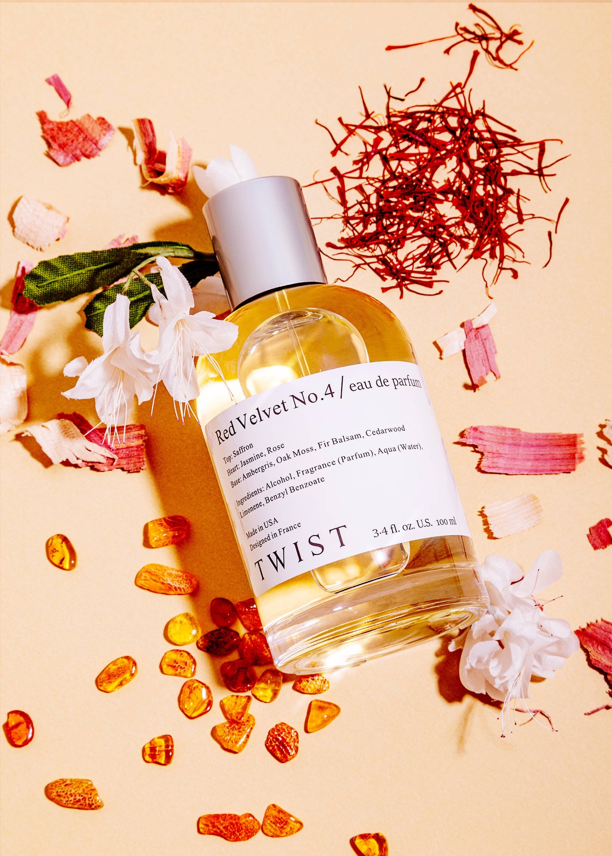 A bottle of Red Velvet No. 4 Perfume from House of Twist rests on a peach background, encircled by white flowers, saffron strands, and amber-hued resin pieces—a striking display for this long-lasting unisex fragrance.