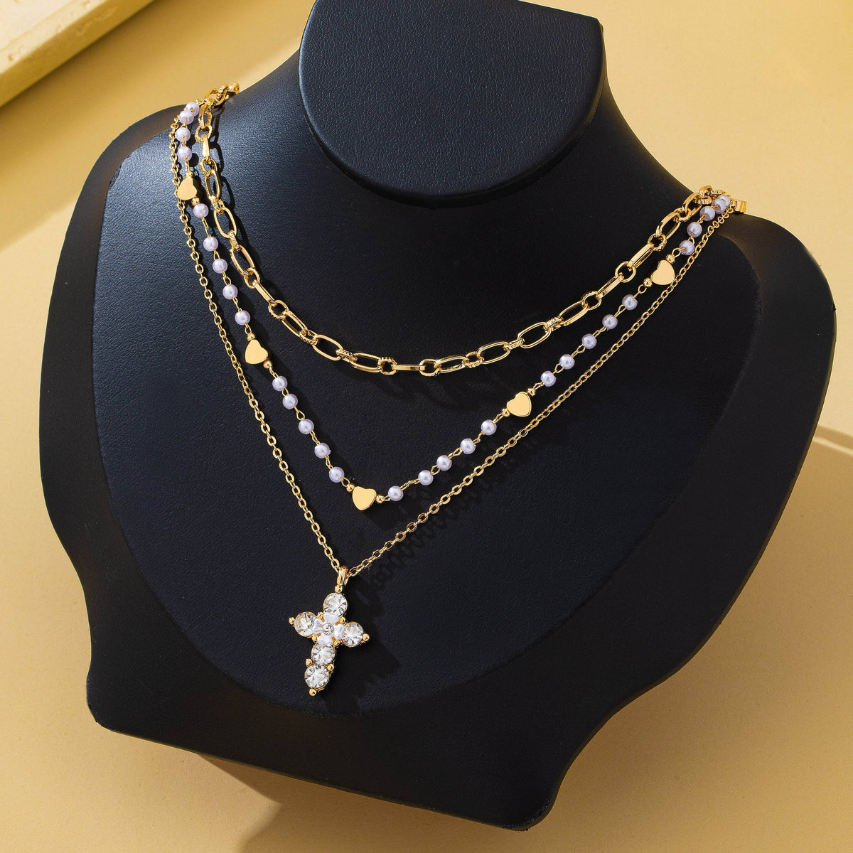 Showcased on a black display bust, the Multi-Strand Pearl & Crystal Cross Necklace by JOSSLYN (wall to wall) features three gold layers: one adorned with small hearts, another strung with white pearls, and a third highlighted by a jeweled cross pendant—all set against a soft beige background.