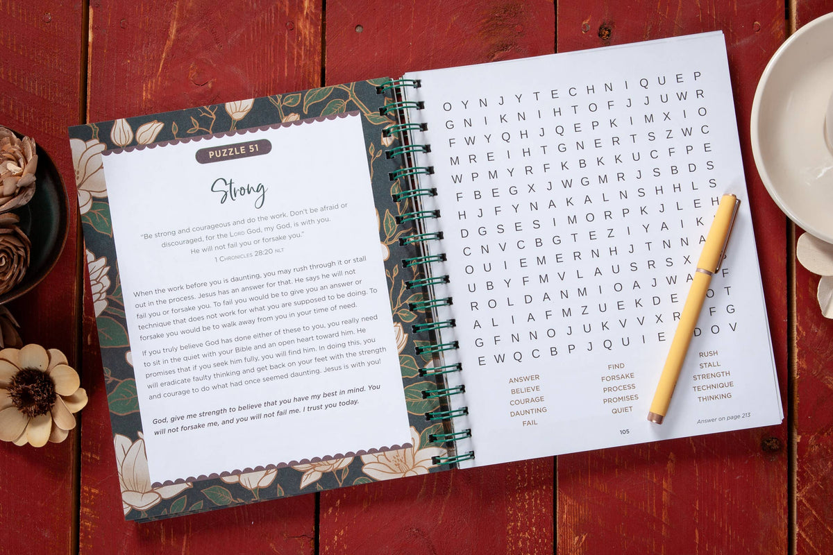 An open copy of the 100 Days of Strength for Women Word Search Book by BroadStreet Publishing Group, LLC features a floral border, with a story on the left page and a word search puzzle on the right. A yellow pen rests atop the book, which is set on a red wooden table next to a cup and some pinecones.