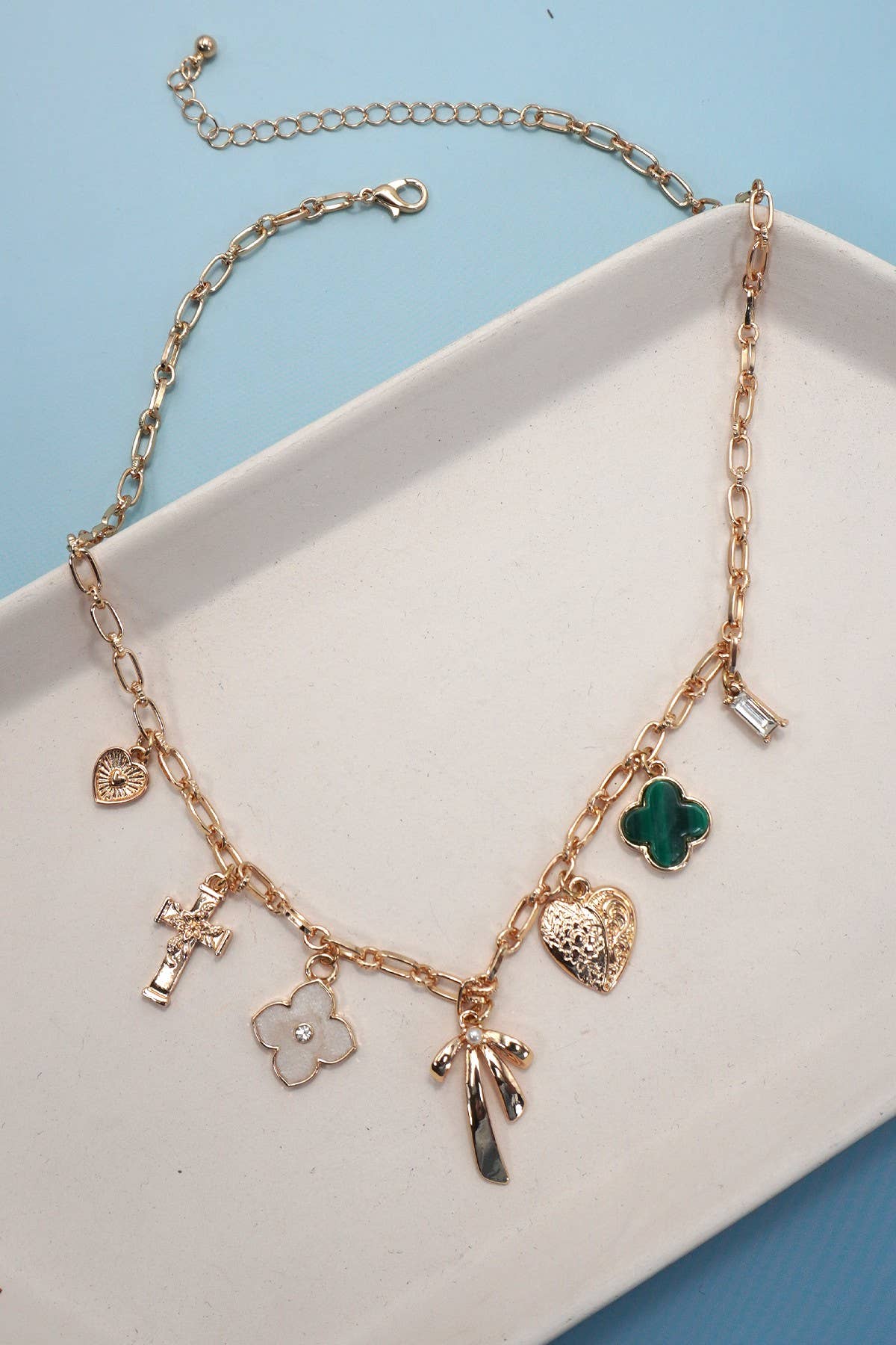 The JOSSLYN by wall to wall Gold Charm Necklace features a gold chain adorned with an array of charms, including a cross, hearts, a green clover, a bar, a white flower, and a hand. The necklace is shown displayed on a white tray set against a blue background.