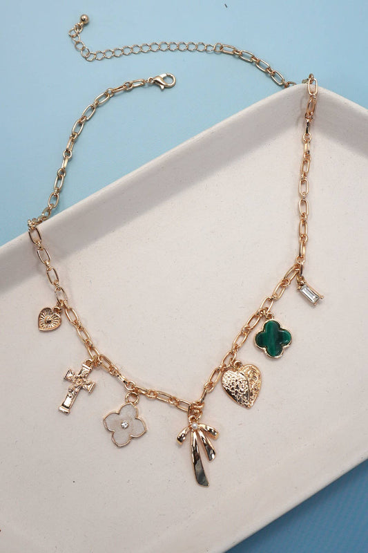 The JOSSLYN by wall to wall Gold Charm Necklace features a gold chain adorned with an array of charms, including a cross, hearts, a green clover, a bar, a white flower, and a hand. The necklace is shown displayed on a white tray set against a blue background.