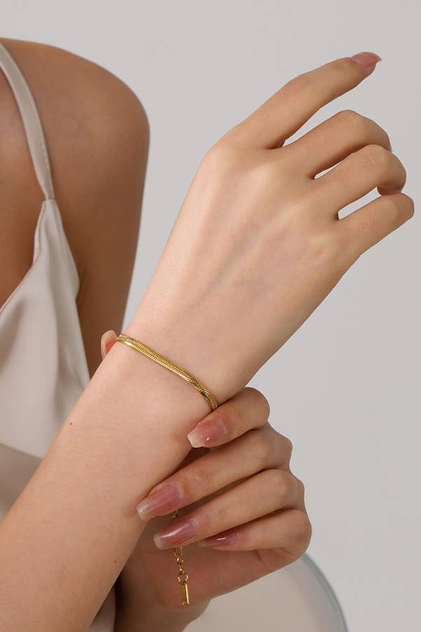 A person with manicured nails wears the JOSSLYN by wall to wall Dainty Gold Bracelet on their wrist. They are dressed in a cream-colored top and gently touch their raised hand with the other, set against a plain, light-colored background.