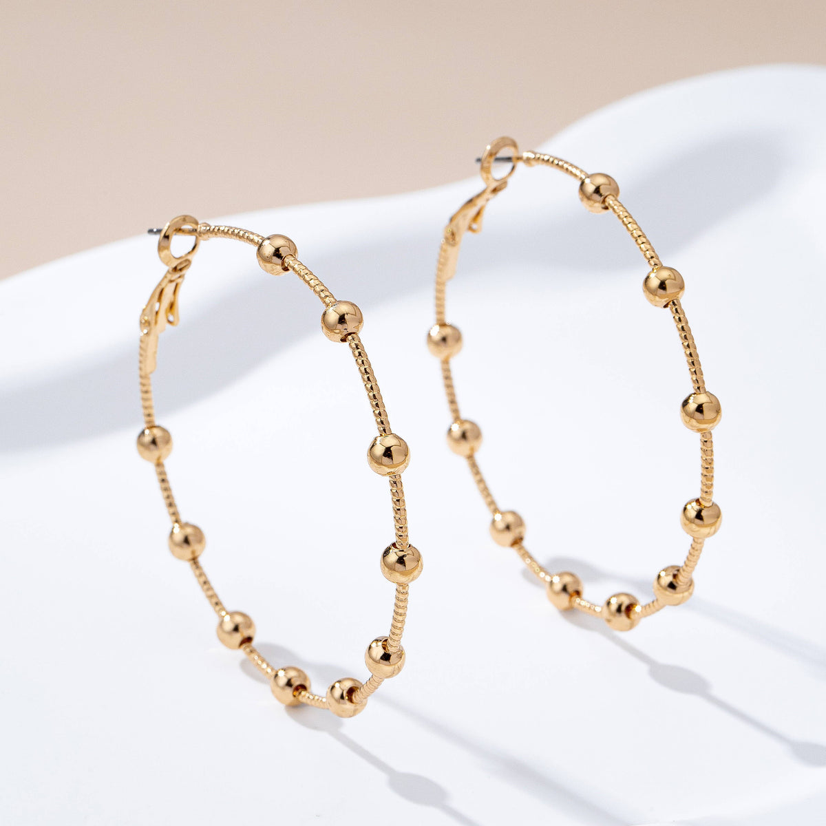 JOSSLYN by wall to wall Gold Ball Beaded Hoop Earrings featuring evenly spaced small gold beads, displayed on a white surface with a soft beige background.