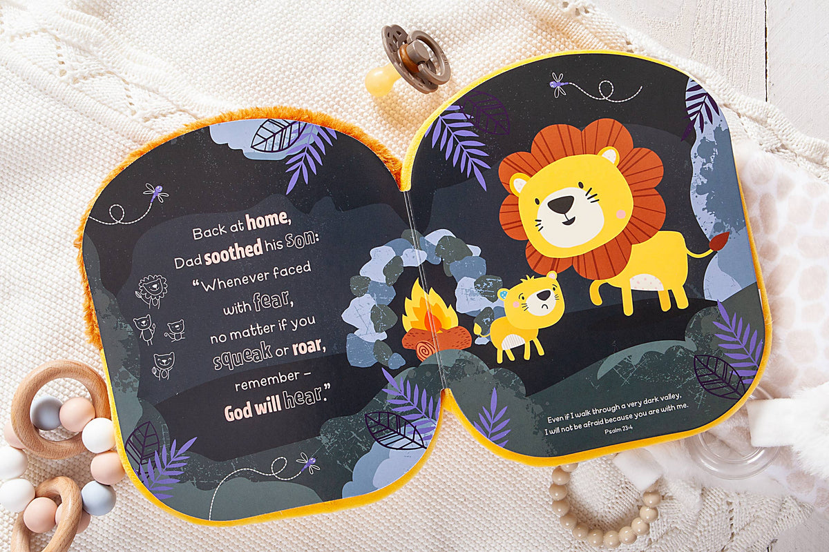 The Squish and Snugg Lion Book by BroadStreet Publishing Group, LLC is open to a page featuring an illustrated lion and cub near a campfire; comforting and reassuring text is displayed on the left page. The book rests on a lightly textured surface, surrounded by pacifiers and baby toys.